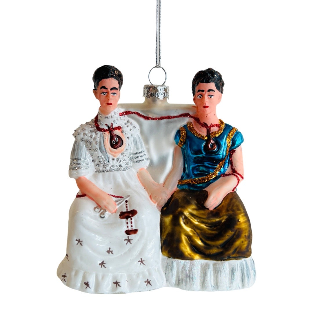 Hand-painted glass two Fridas ornament with glitter accents. The ornament captures the real life oil painting of Frida Kahlo called, "The Two Fridas." It is a double self-portrait, depicting two versions of Kahlo seated together.