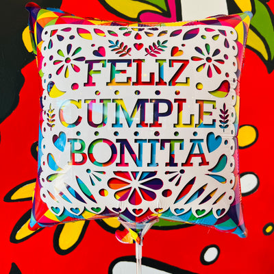 "Feliz Cumple Bonita" phrase square balloon pictured in front of Artelexia Frida mural. Design features white papel picado with rainbow background.