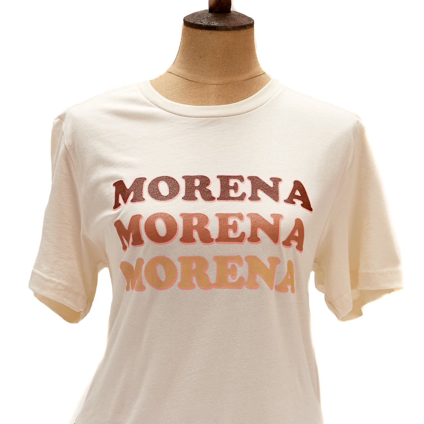 Light tan, "Morena" t-shirt pictured on mannequin. T-shirt features three different shades of brown with pink outline on phrase.