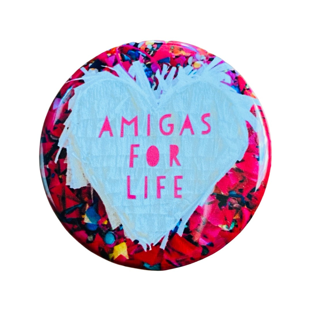 Round pin back button with a confetti back ground and features a white pinata heart with the phrase Amigas for Life.
