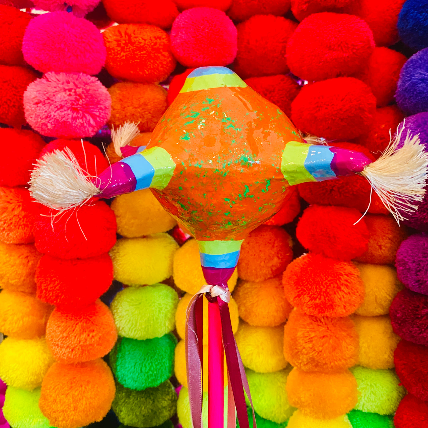 Piñata Paper Mache Shaker with colorful ribbons (Style 1) pictured in front of multicolored pompoms. 