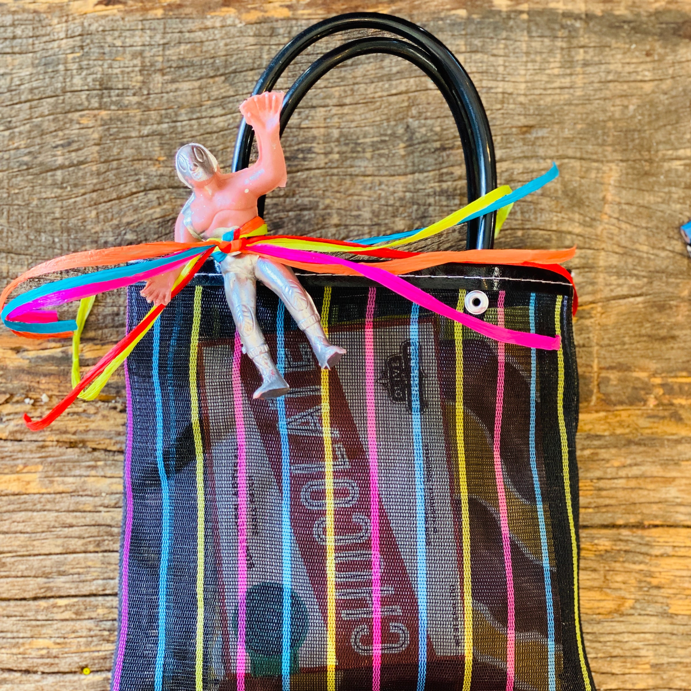 Black woven Mexican mercado bag filled with assorted Mexican candies and stickers. Handles tied off with colorful ribbon and secured with plastic Luchador figurine.
