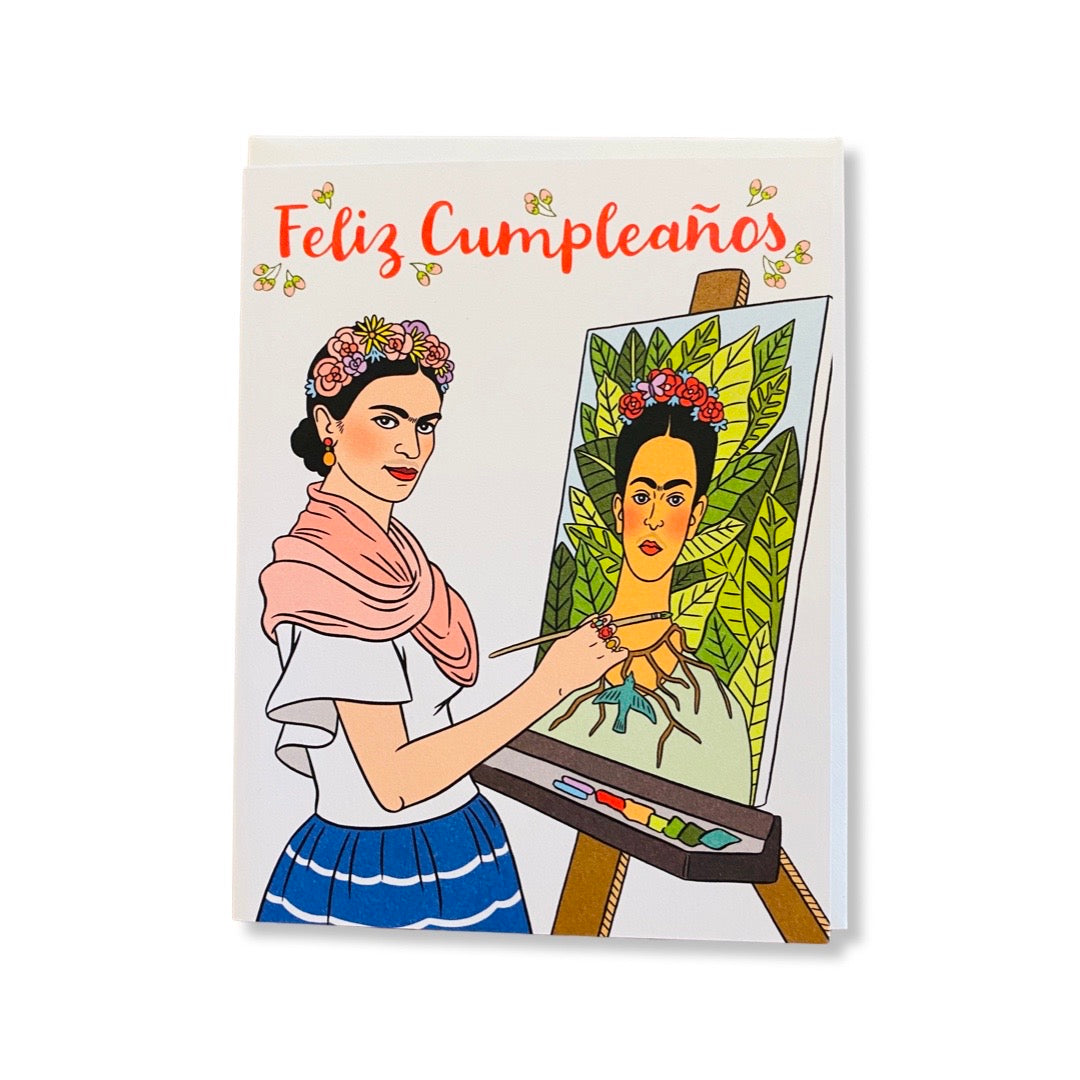 White Feliz Cumpleanos greeting card. Design features Frida Kahlo standing in front of an easel, painting a self portrait.