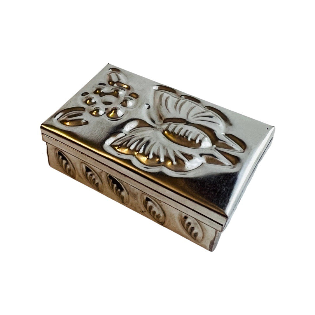 A tin trinket box with a hammered butterfly and flower design on the lid and leaves on the side.