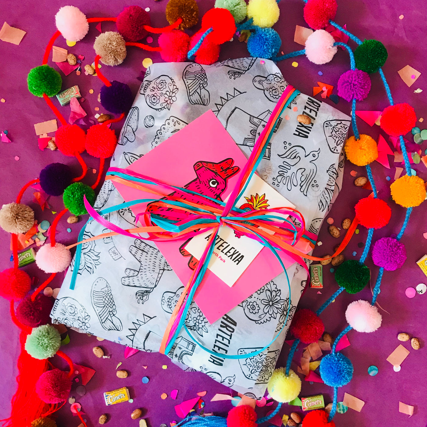 Top view of $75 goodie bag wrapped in Artelexia branded tissue paper and tied with colorful ribbon. Wrapped item includes postcard, business card, sticker, and confetti with beans and gum. Item pictured with colorful pom pom garland on top of pink tissue paper.