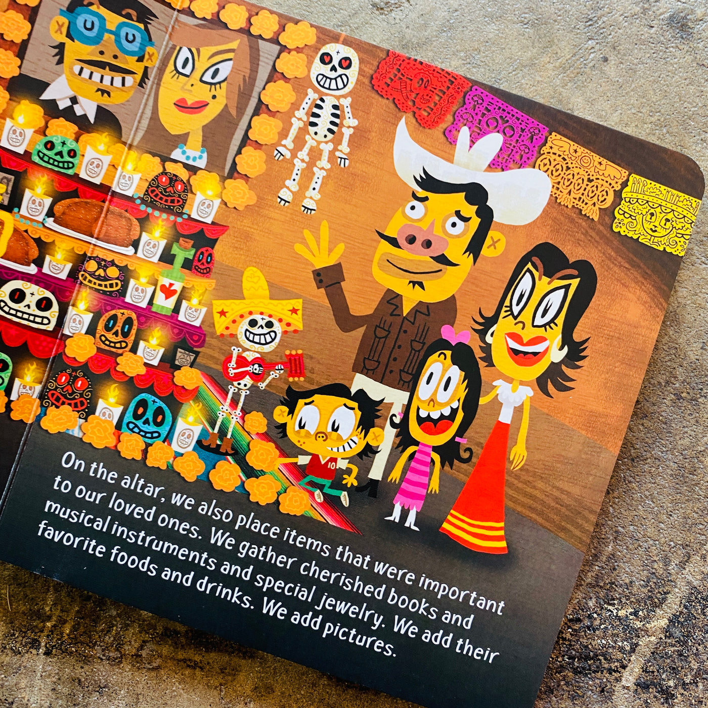 Inside of view of the book featuring a family standing in front of a Dia de los muertos altar with lit candles, colorful papel picado and skeletons playing a guitar.