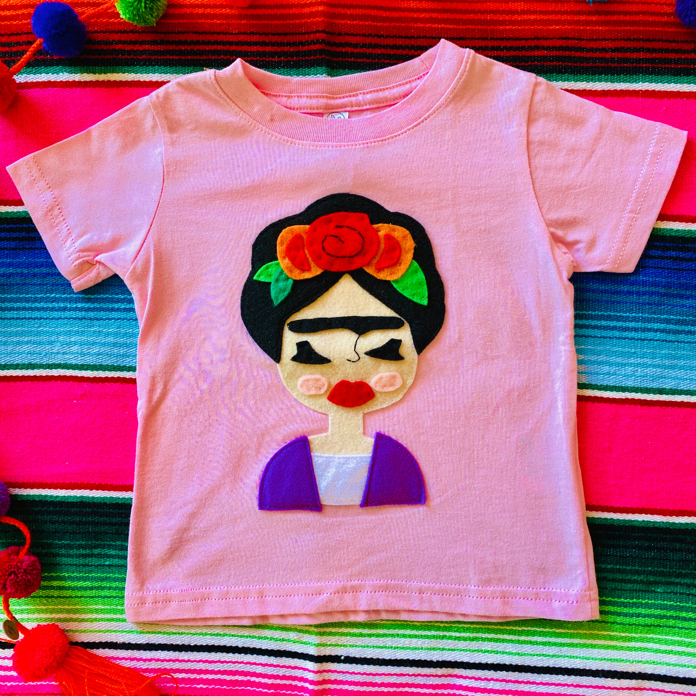 Top view of hand-stitched Frida kid's pink t-shirt. T-shirt pictured on top of serape blanket.