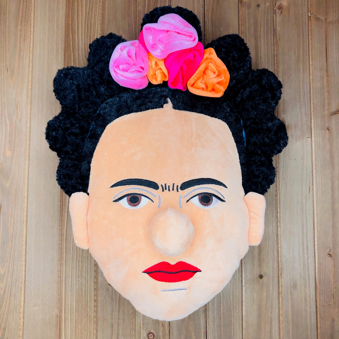Plush Frida Kahlo head pillow pictured on wooden table.