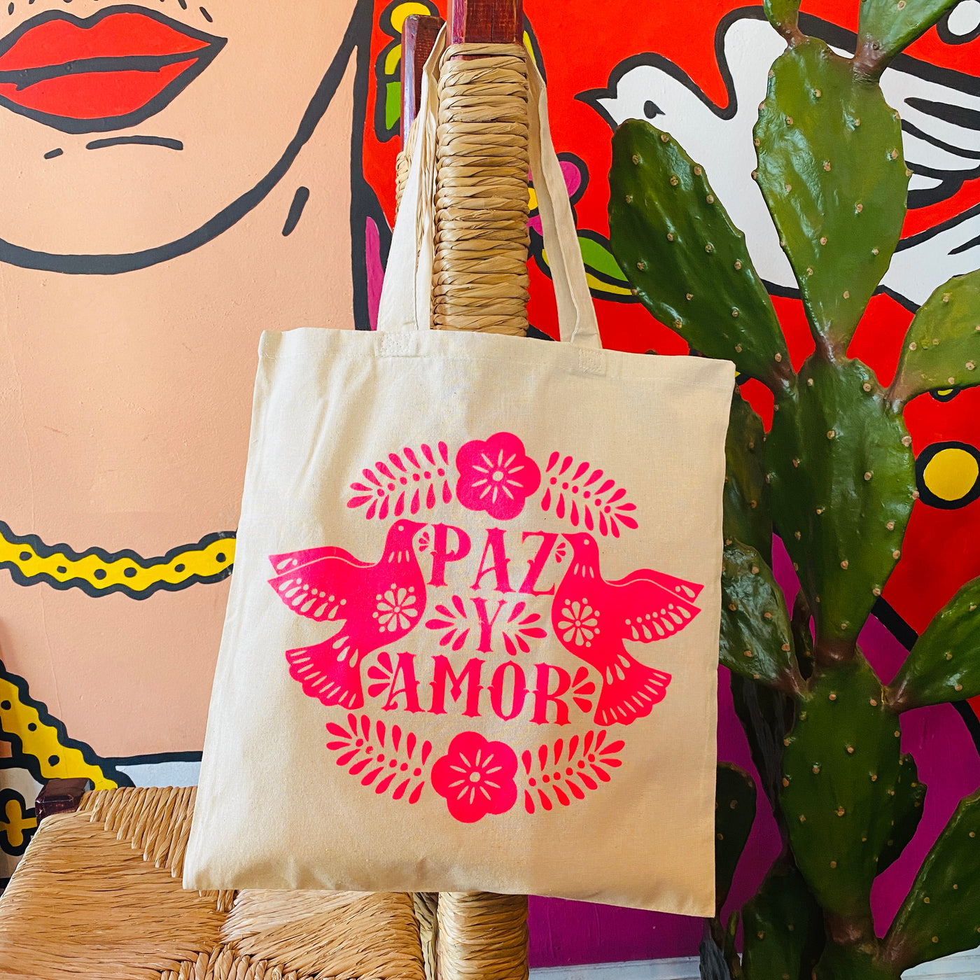 Natural colored tote bag with bright pink design of two doves and the words "Paz y amor" in the center.
