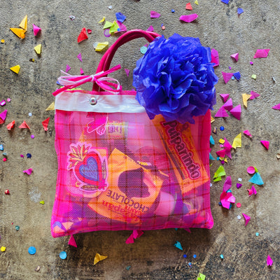 Pink Mexican mercado bag filled with various Mexican candies and stickers. Handles of the bag are tied with colorful ribbon and secured with a blue paper flower.