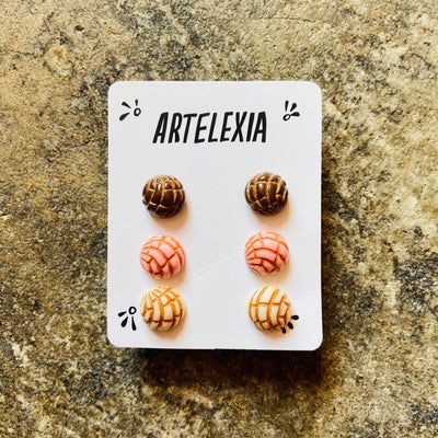 Trio of concha (type of sweet bread) stud earrings in brown, pink, and white with Artelexia branded packaging.  
