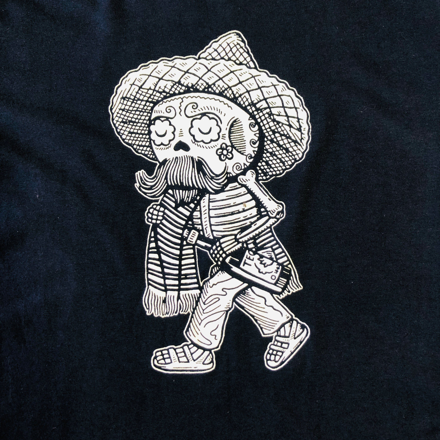 Close up of Men's Borracho Calavera graphic. T-shirt is navy blue with white accents. Design features skeleton with sombrero, serape, and a bottle of tequila.