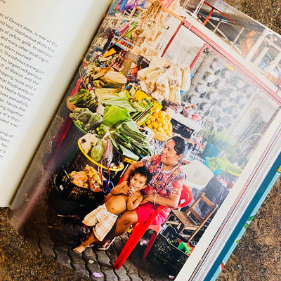 Top view of page inside The Food of Oaxaca: Recipes and Stories from Mexico's Culinary Capital cookbook. Photo features woman sitting with a young child in a mercado surrounded by different food items.