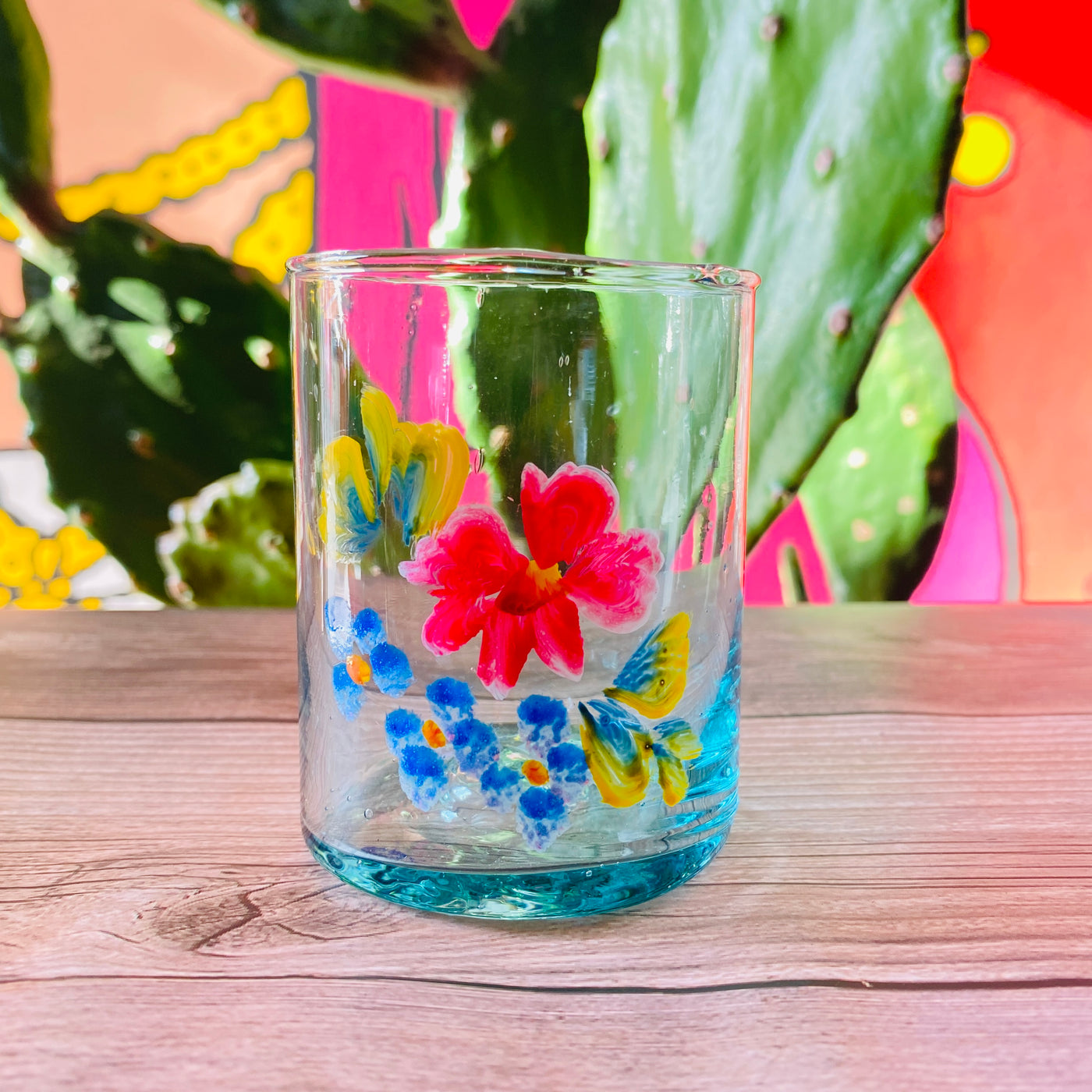 Hand-painted flower glass pictured on top of wooden table with cactus in the background.