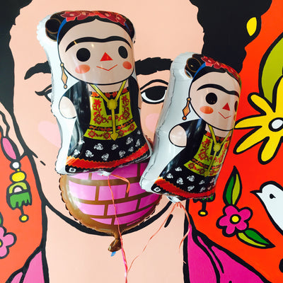 Two Frida Kahlo balloons floating next to each other inside of Artelexia store and in front of "Frida" mural. Frida has a Maria doll style appearance. Main colors featured are red, black, and yellow. Frida wears a red flower crown and traditional dress