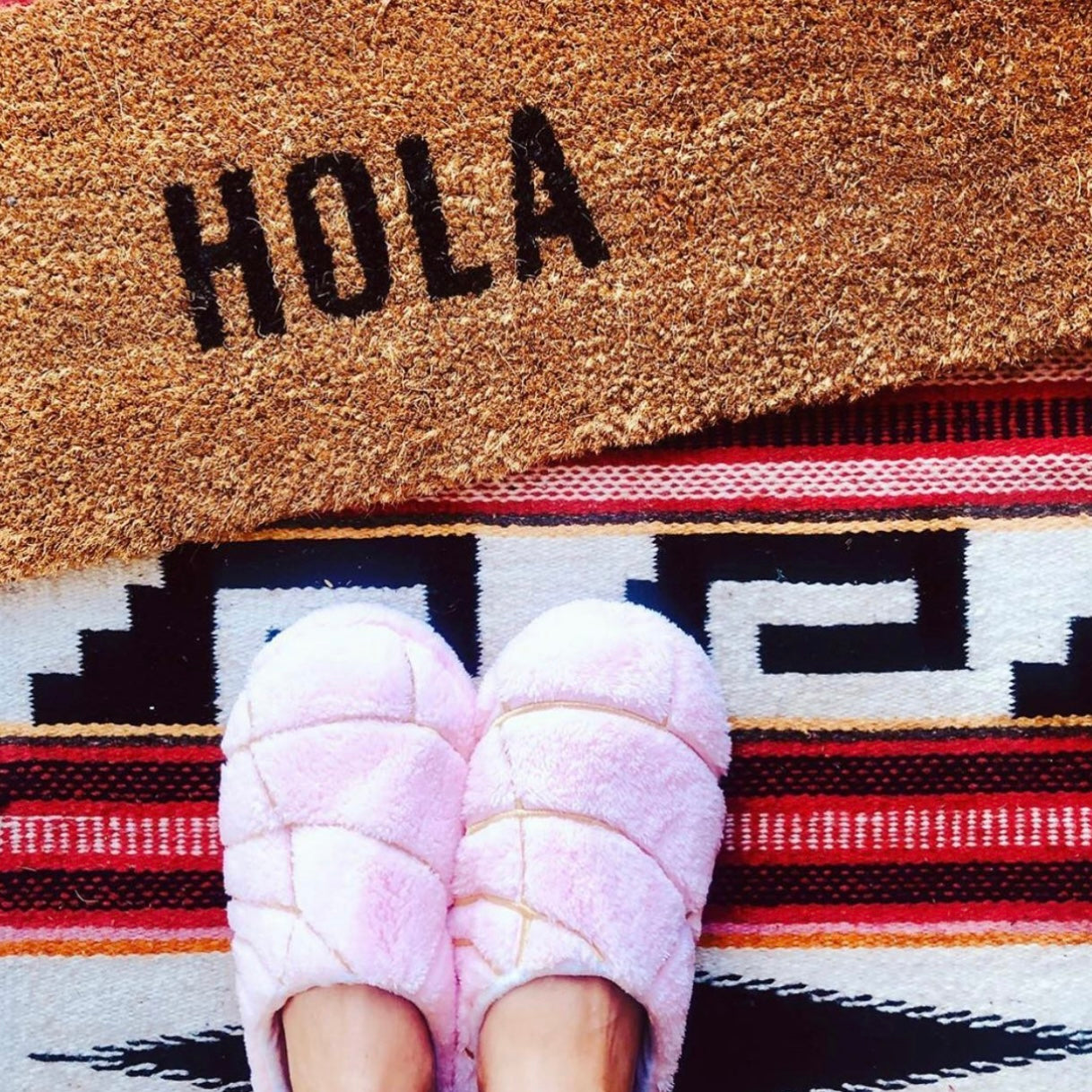 Top view of light pink concha pantuflas (slippers). Photo features HOLA welcome mat.