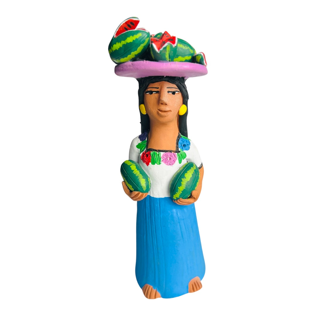 Ceramic figurine that features a woman in a white top and blue skirt holdig two watermelons and a tray of watermelons on her head.
