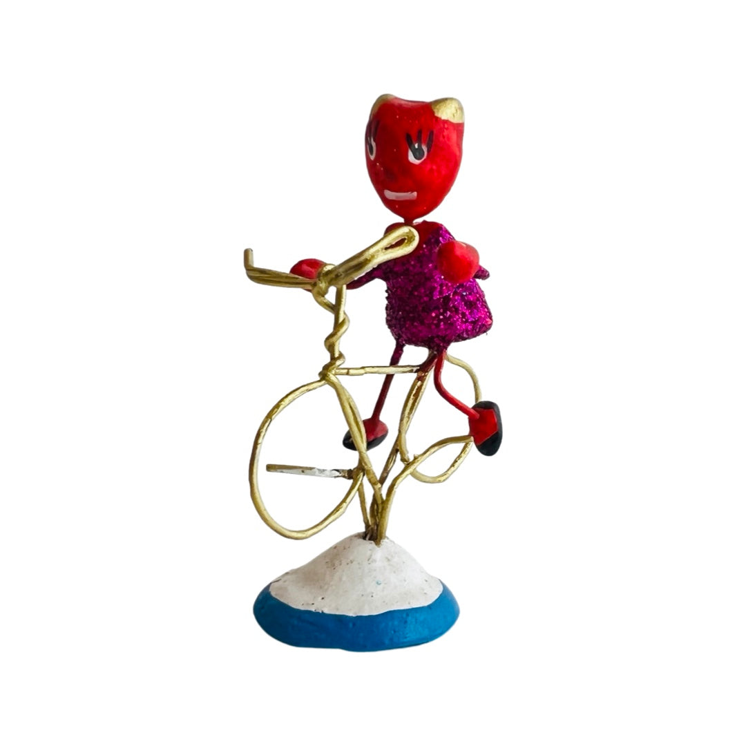 Side view of a painted clay devil with a glittery torso on a bike made of wire. 