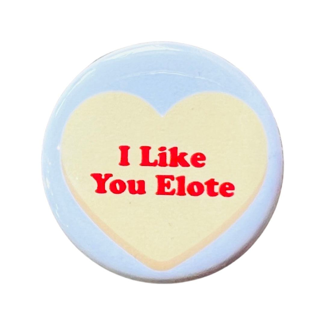 White round pin back pin featuring a yellow heart and the phrase I Like You Elote in red lettering.