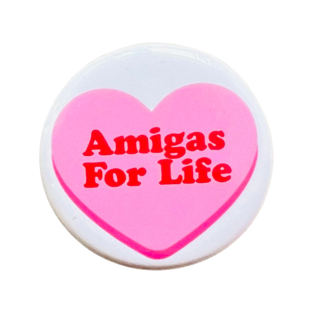 Round white pin back button featuring a pink heart and the phrase Amigas For Life in red lettering.