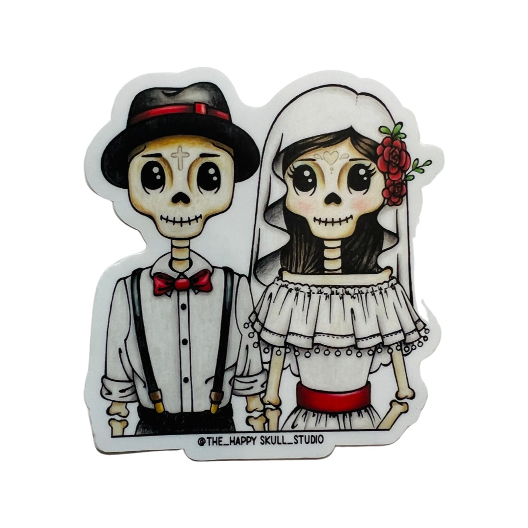 A skeleton groom and bride with the groom wearing a black hat and suspenders and the bride wearing a veil with red flowers on one side as well as a red belt.